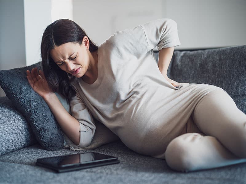 Pregnant woman experiencing back pain on sofa with tablet nearby.